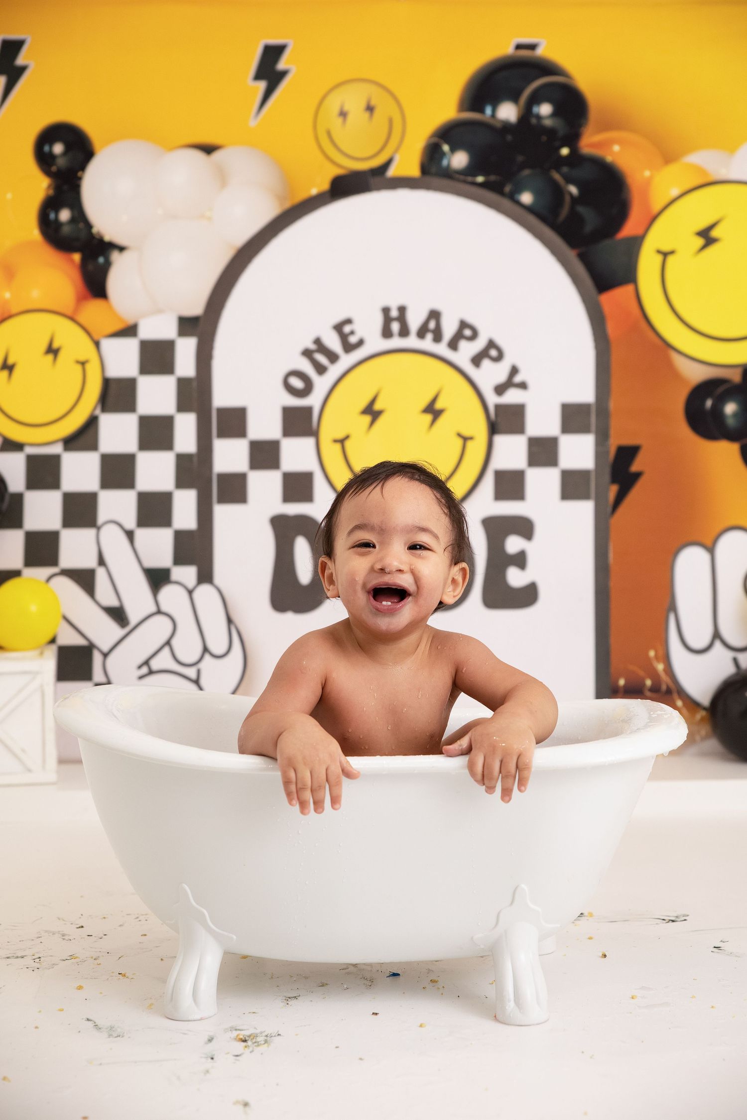 A laughing baby sits in a white mini bathtub in front of a colorful smiley face balloon backdrop for a first birthday.
