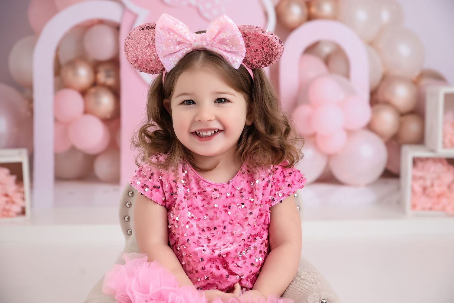 A smiling little girl in a pink sequin dress and Minnie Mouse ears poses in front of pink balloon decorations.