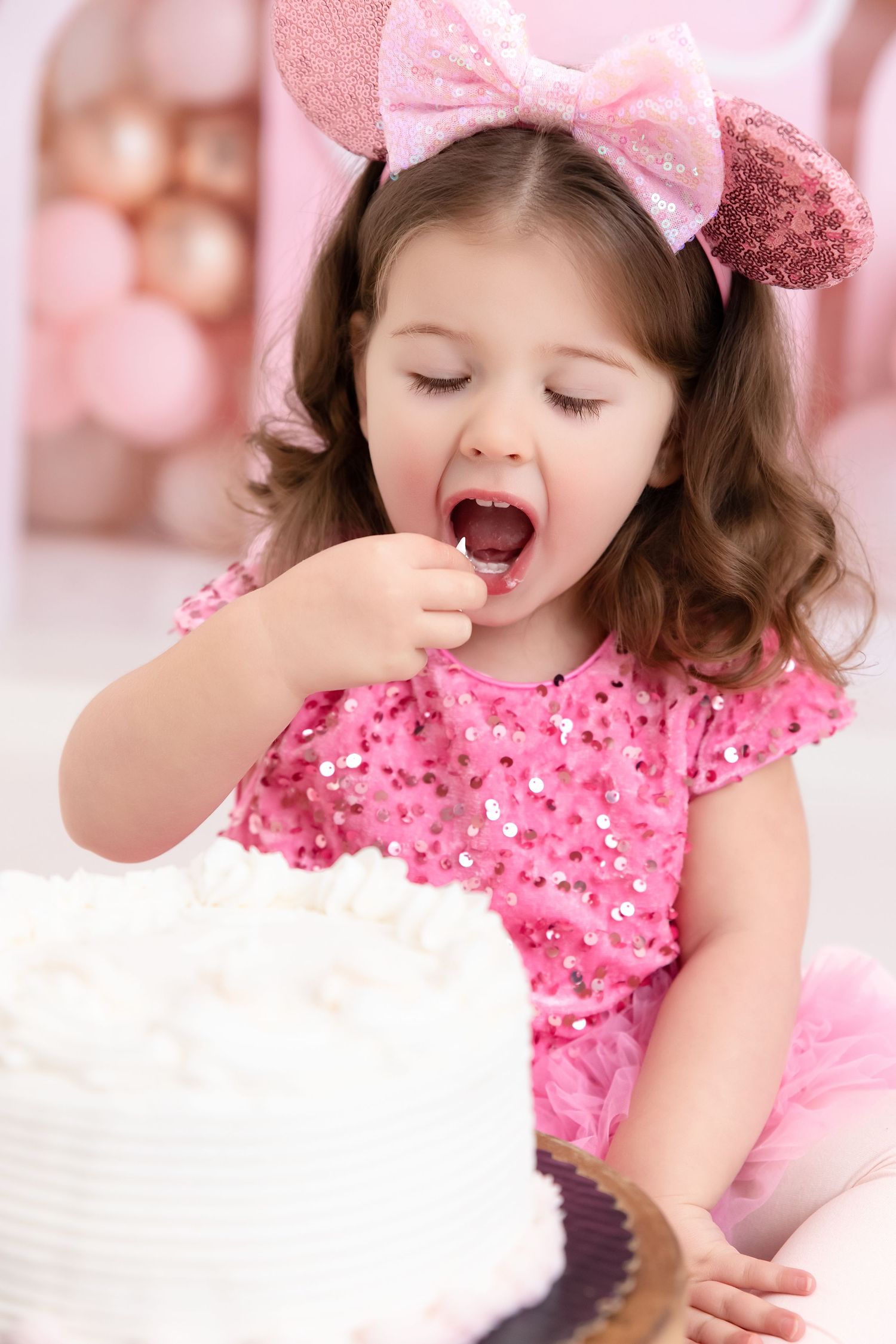 A cute little girl in a pink sequin dress and Minnie Mouse ears eagerly eating her white birthday cake.