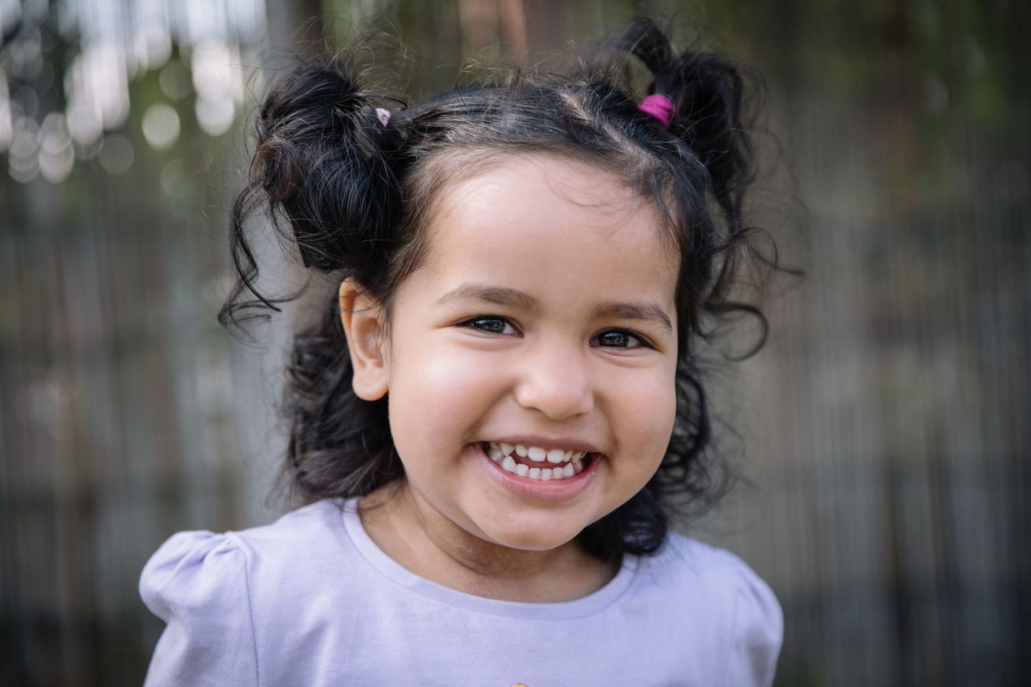 A child wearing a lavender colored shirt smiles brightly with dark curly hair in pigtails against a blurred background.