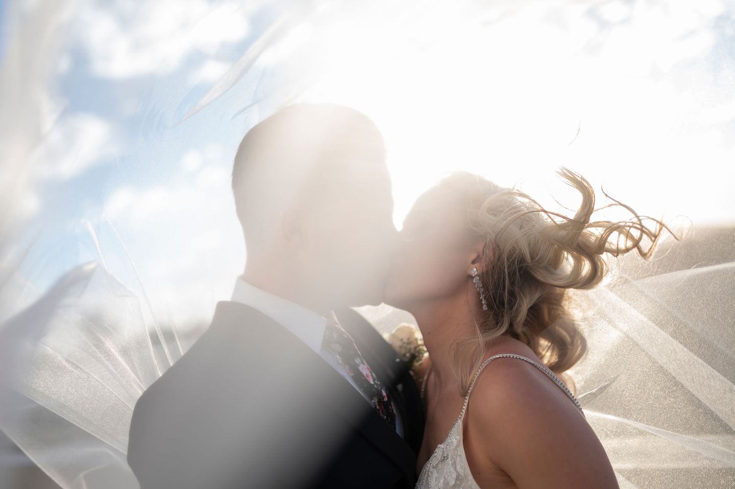 Bride and groom sharing a romantic kiss under a flowing veil with sunlight and blue sky in the background.