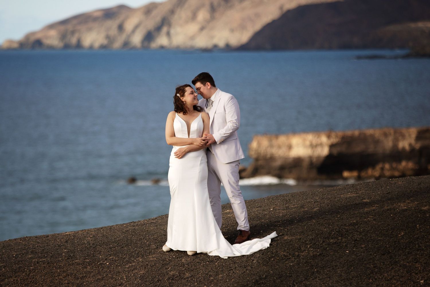 Bride and groom embrace on rocky beach of La Pared in Fuerteventura. Ocean and cliffs backdrop, bride in white gown, groom in light suit.