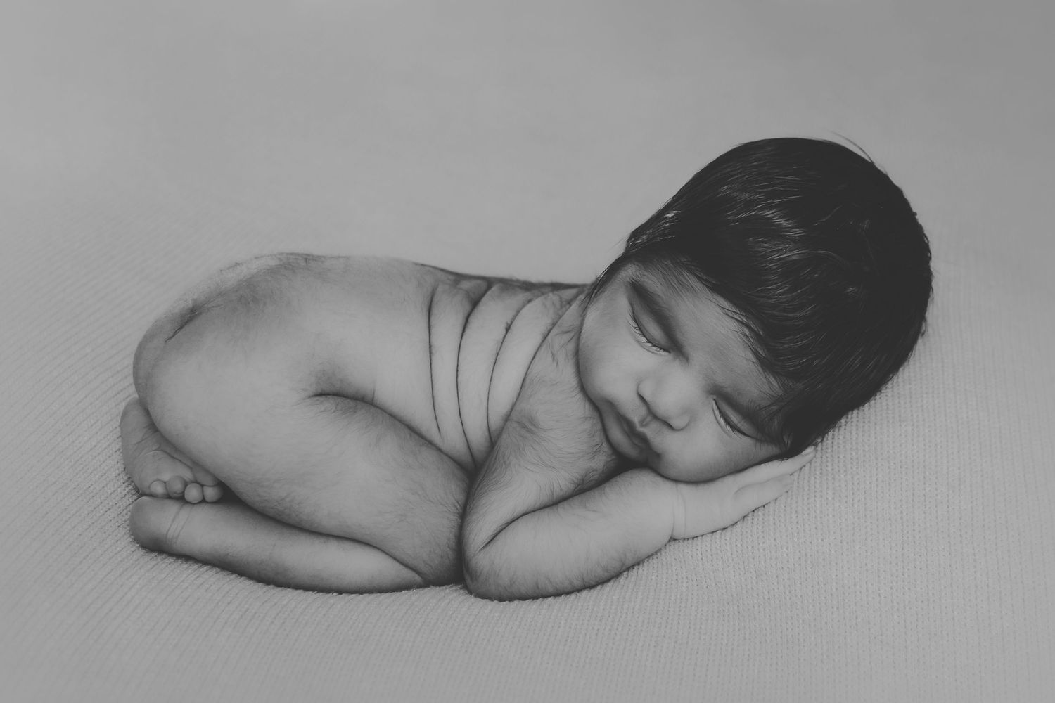 Black and white photograph of a sleeping newborn baby lying on a white blanket in a peaceful pose.