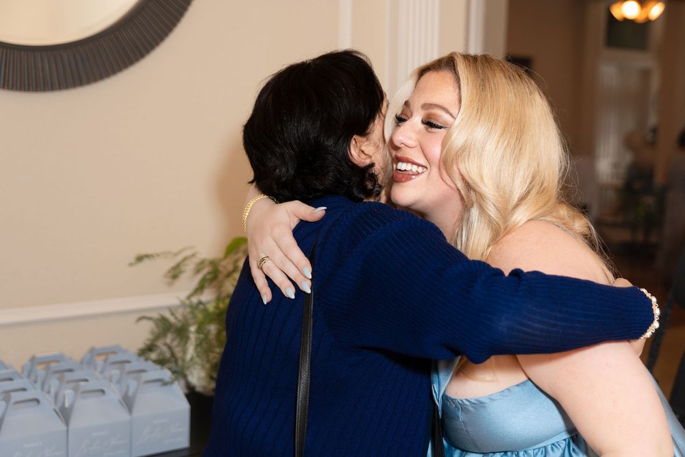 Two people share a warm embrace at an indoor event, one wearing a navy sweater and the other in a light blue dress.