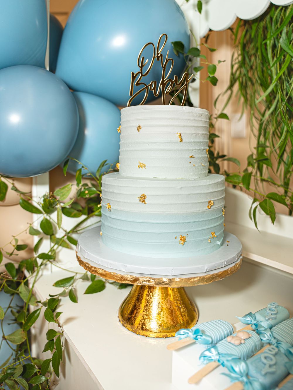 White wedding cake with gold accents and blue balloons displayed on a gold stand with greenery decorations.