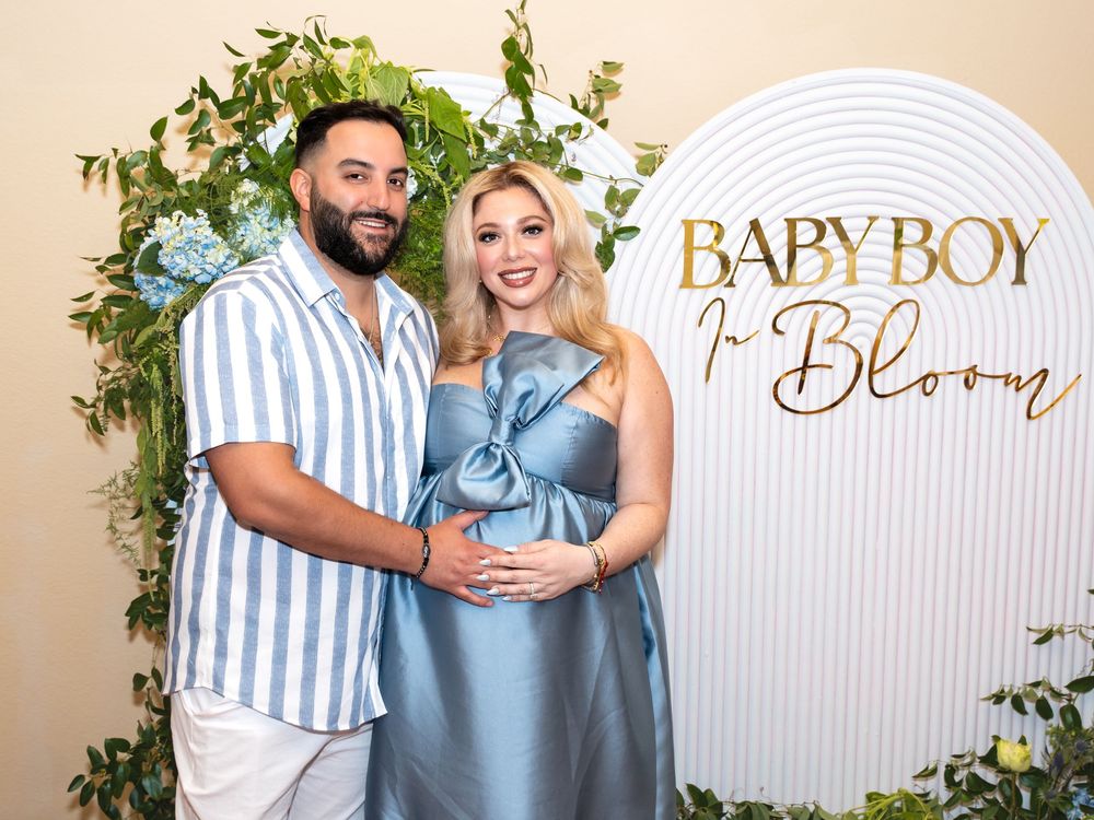 Couple in coordinated blue and white outfits pose together at a baby shower celebration with decorative floral backdrop.