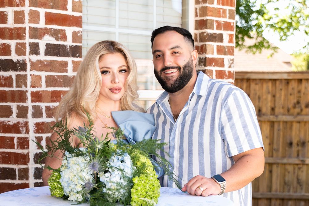 A couple poses together in front of a brick wall with white and green floral arrangements in a casual outdoor setting.