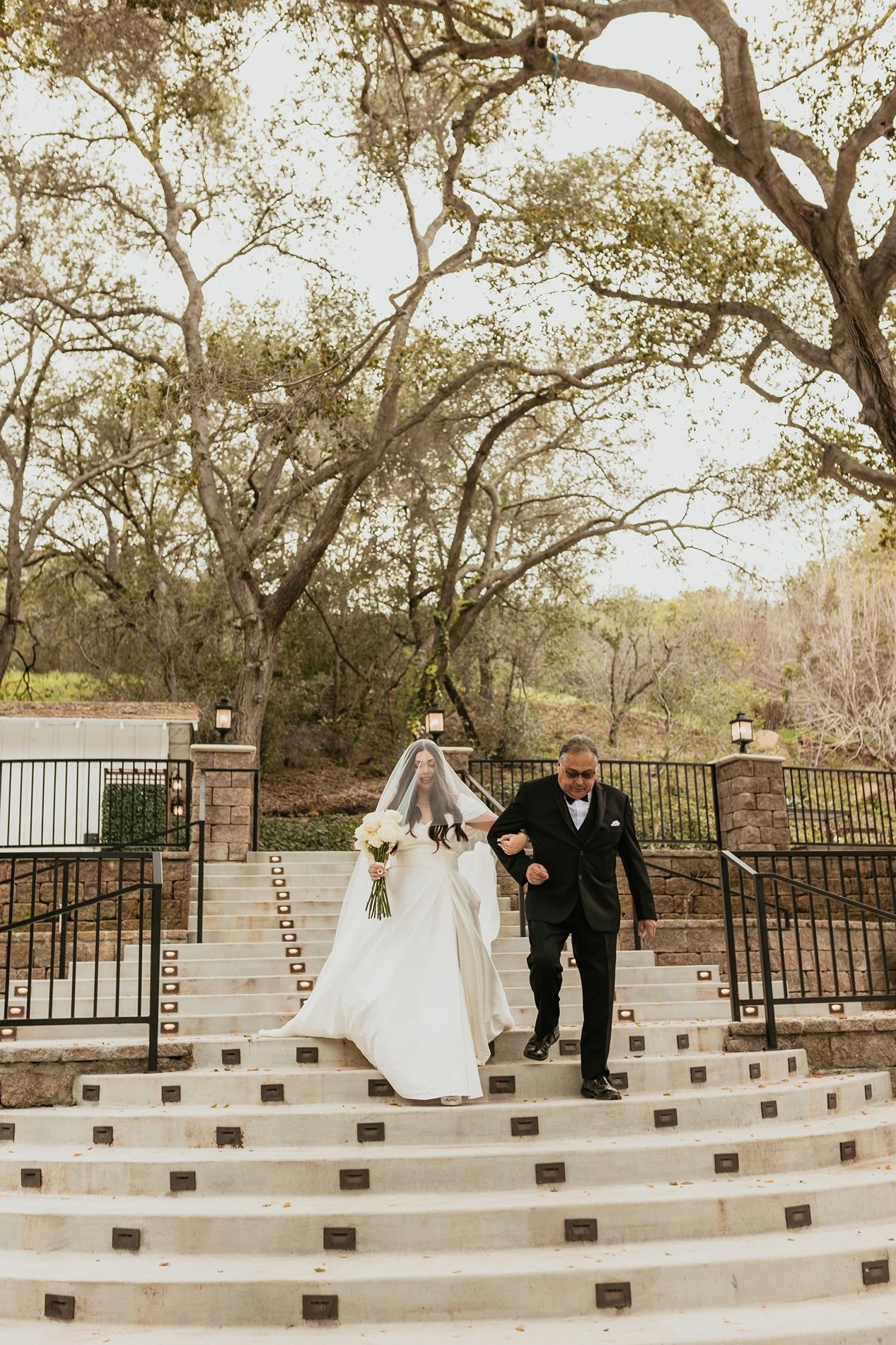 A bride in white dress walks down curved outdoor stone steps with an escort under arching trees at The Oaks at Duncan Lane.