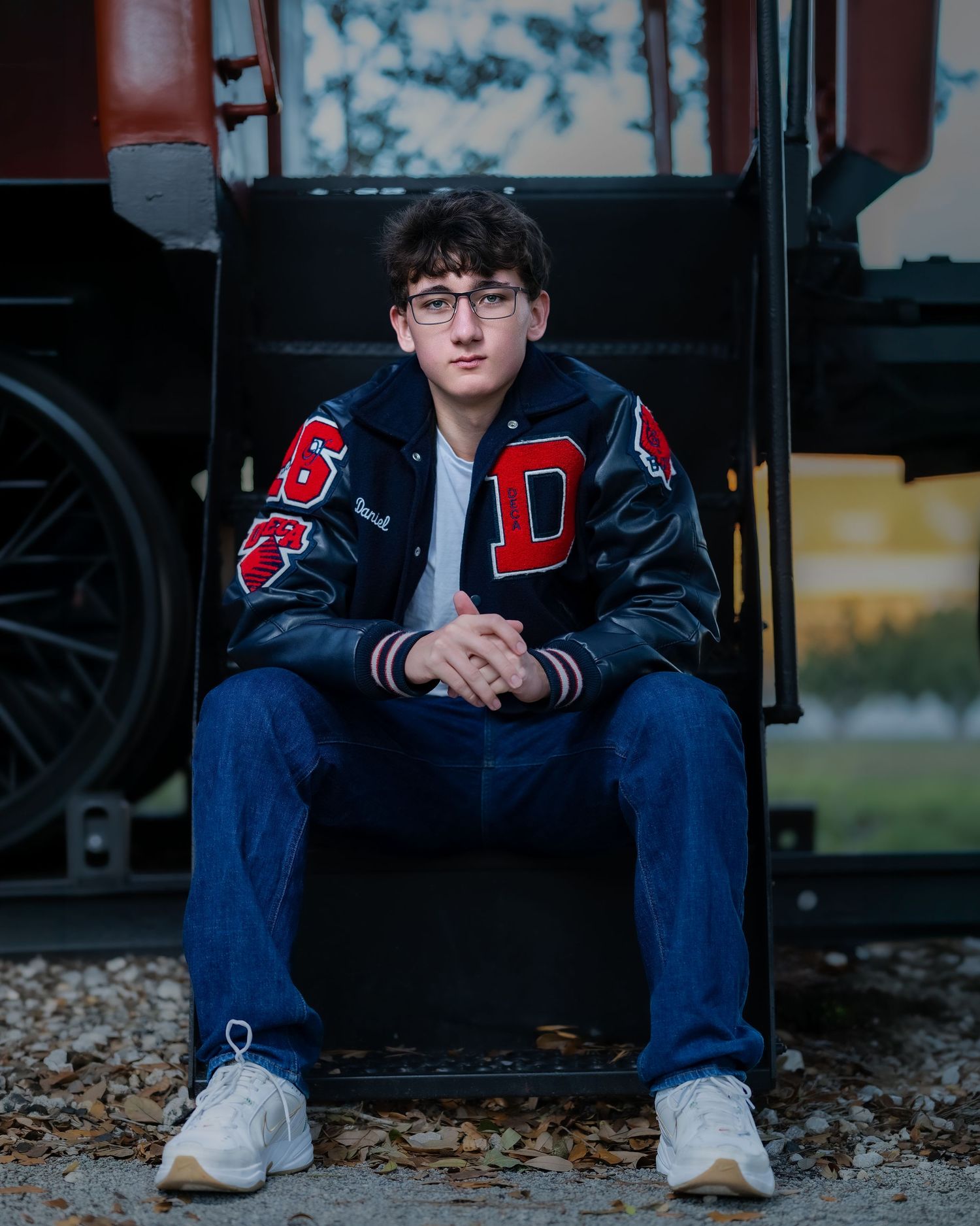 Student in varsity letterman jacket sits on school bus steps during autumn with fallen leaves on ground.