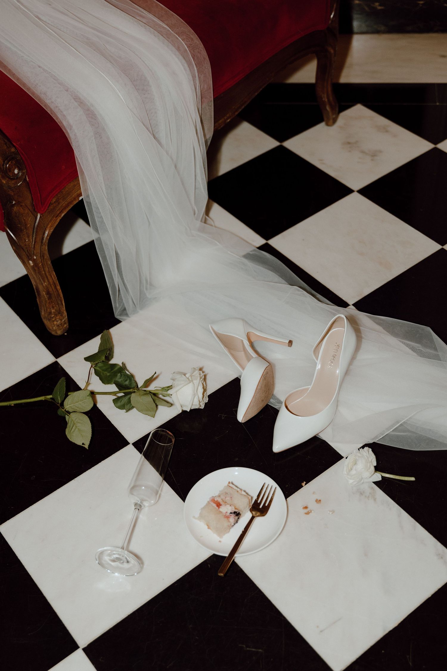 White wedding heels, veil, and floral details arranged on black and white checkered floor with champagne glass and dessert plate.