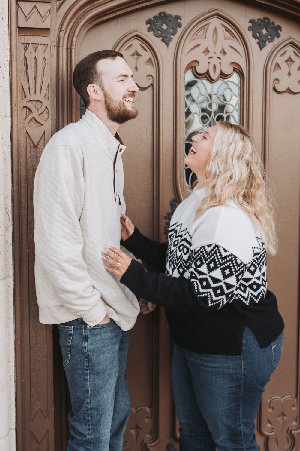 A couple wearing winter sweaters share a laugh while standing in front of an ornate wooden door with Gothic details.