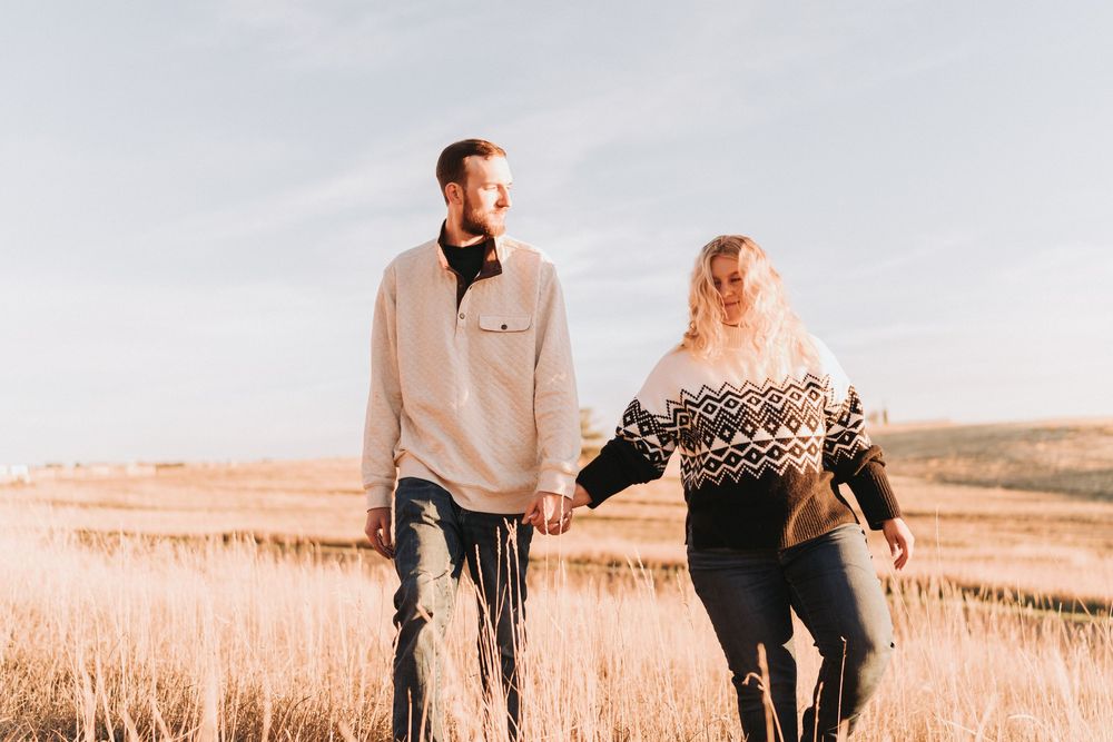 A couple walks hand in hand through a golden wheat field at sunset in warm autumn light.