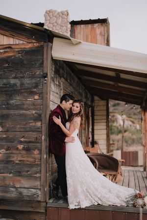 Bride and groom embracing on rustic wooden cabin porch, bride in lace gown, groom in burgundy velvet suit.