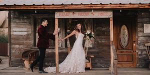 Couple poses at rustic wooden venue entrance, bride in lace gown, groom in burgundy suit, boho wedding style.