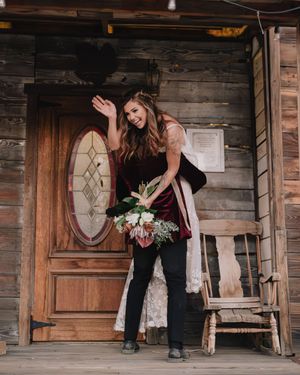 Smiling bride in burgundy velvet dress holding white bouquet, waving on rustic wooden cabin porch.