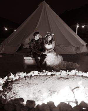 Couple sitting together by a glowing campfire in front of a bell tent at night, black and white wedding photo.