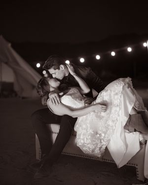 Black and white photo of a couple sharing a romantic dip kiss in wedding attire under string lights at night.