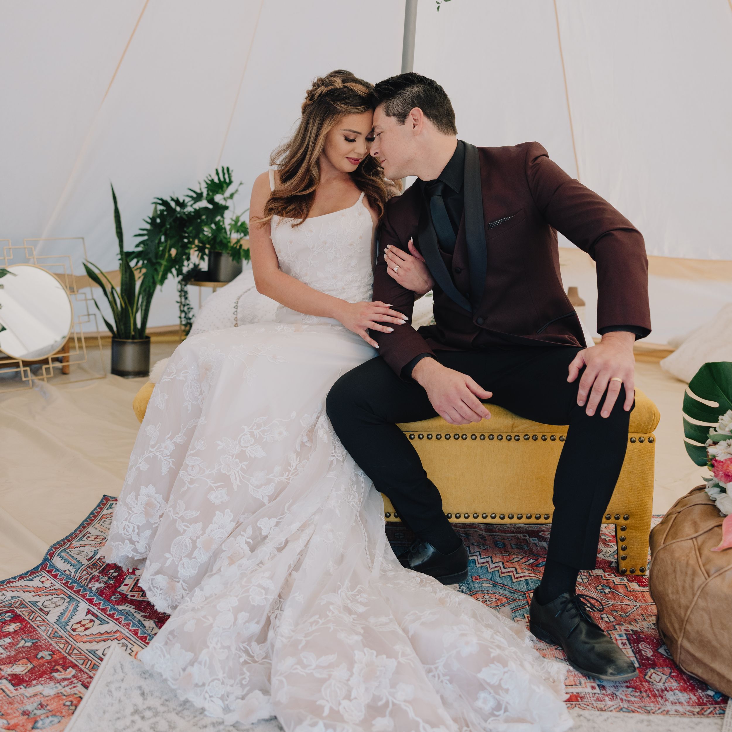 Bride in lace gown and groom in burgundy blazer sit intimately on a yellow sofa inside a boho tent.