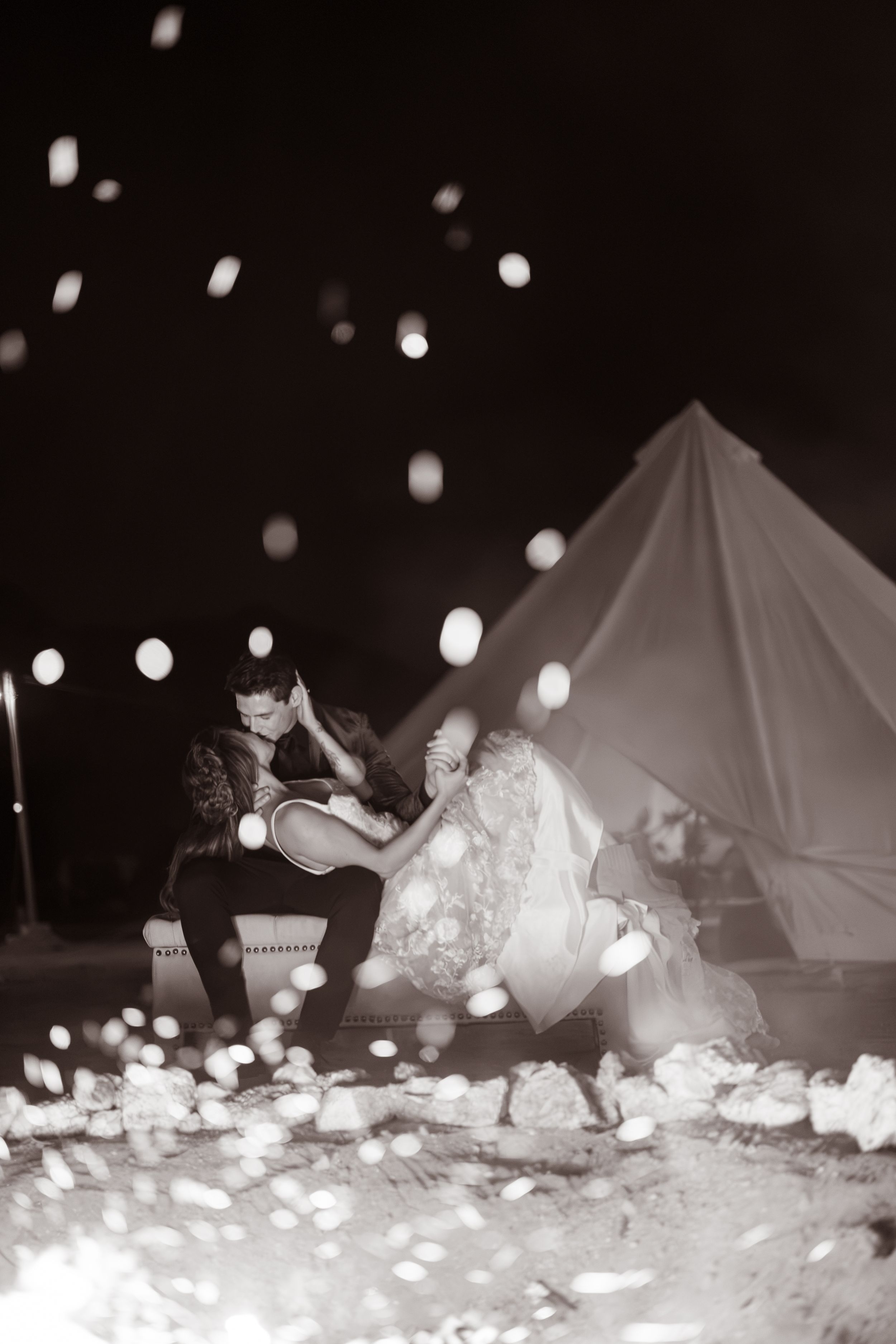 Black and white photo of a bride and groom sitting together outside a glowing tent at night with bokeh lights.