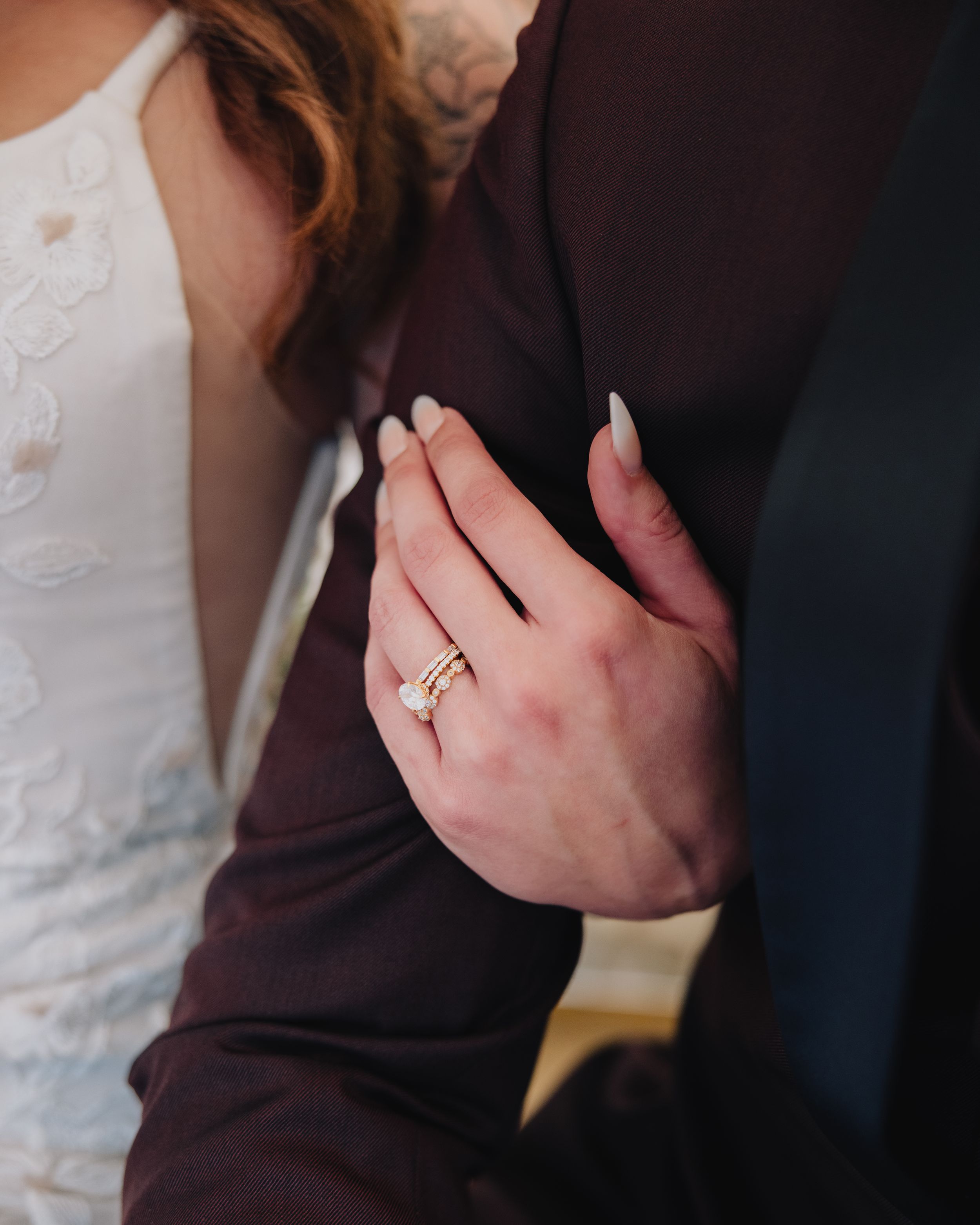 Close-up of a bride's hand with a gold diamond engagement ring, resting on groom's dark suit sleeve.