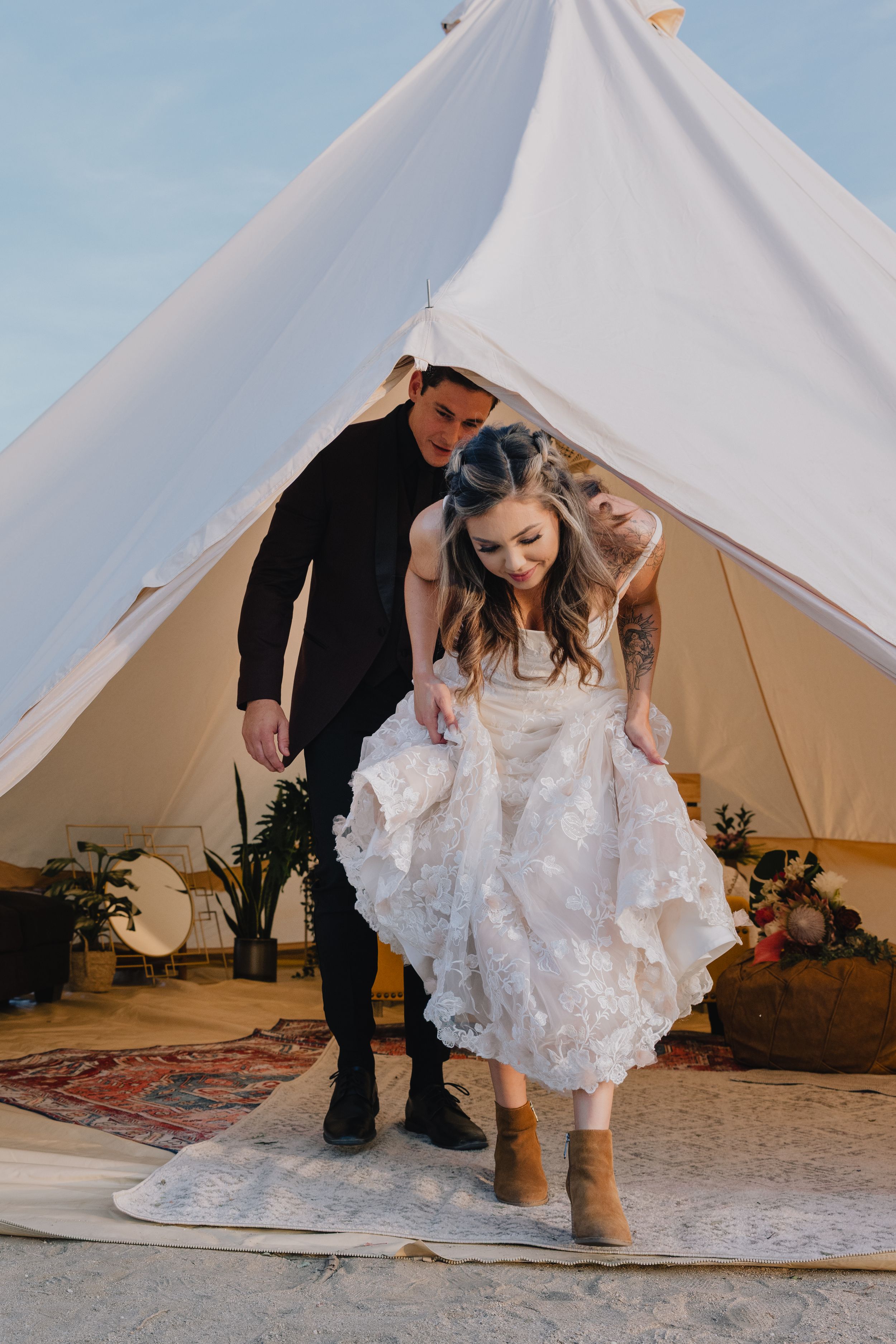 Bride in ruffled white dress and boots exits a glamping bell tent with groom in black suit and bridesmaid.