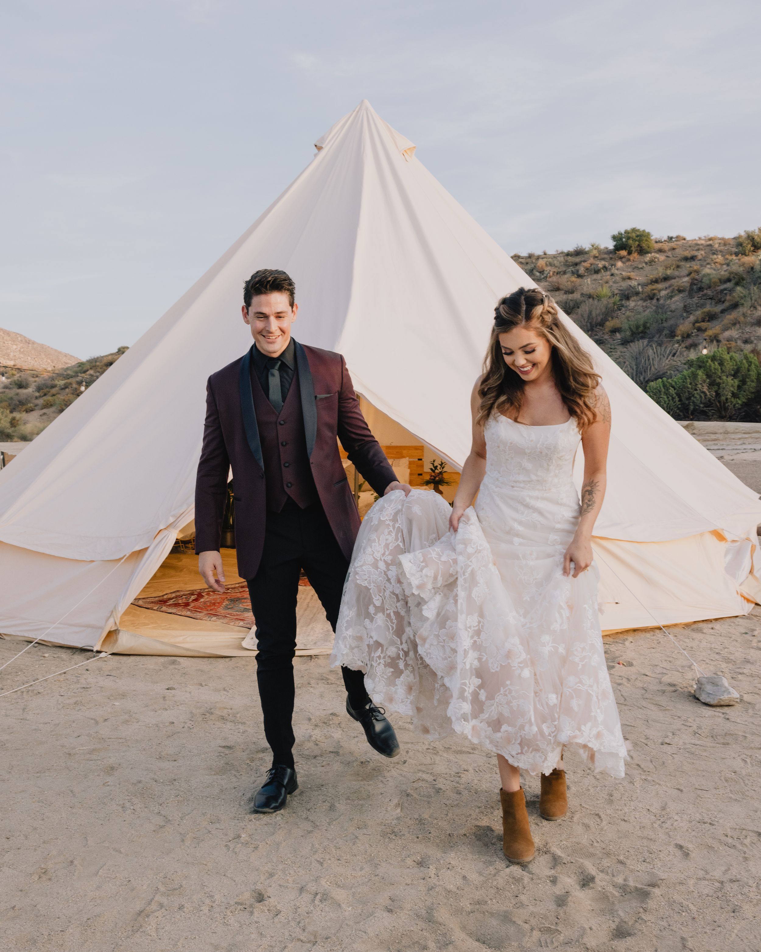Bride and groom walking hand in hand outside a white bell tent on sandy terrain.