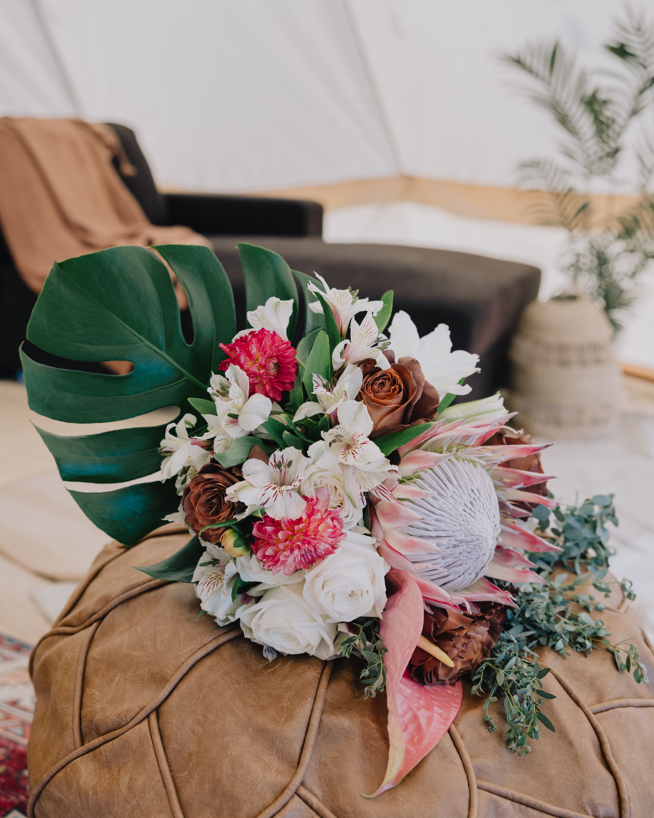 Tropical wedding bouquet with monstera leaf, protea, carnations, and white blooms on leather pouf.