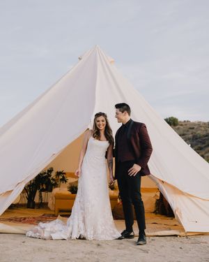 Bride in lace gown and groom in burgundy tuxedo hold hands before a white bell tent on a sandy beach.