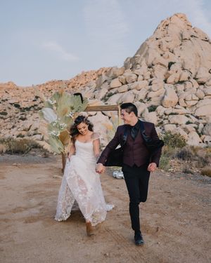 Bride in lace gown and groom in burgundy suit walking hand-in-hand in a rocky desert landscape.