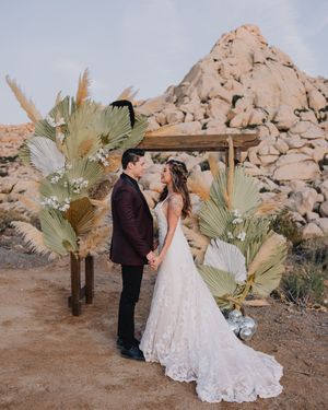 Couple shares a romantic moment at a boho desert wedding altar with pampas grass and dried palm leaves, rocky landscape behind.