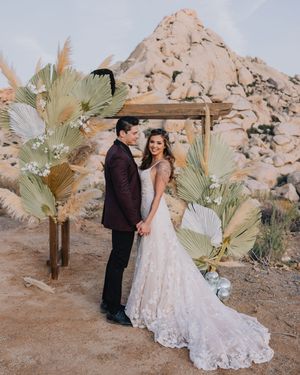 Couple exchanges vows under rustic wooden arch adorned with dried palms and pampas grass in Joshua Tree desert.