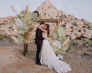 Couple shares first kiss under rustic wooden arch adorned with pampas grass and palm leaves in desert wedding.