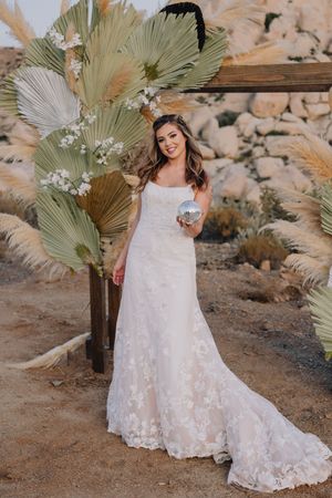 Bride in floral lace wedding gown holding a disco ball near palm fronds in a desert setting.