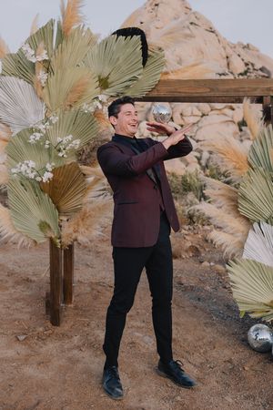 Groom in burgundy blazer poses playfully with disco ball near boho desert wedding arch with dried palm decor.