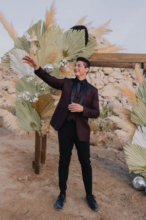 Groom in burgundy velvet blazer poses with disco ball amid dried palm leaf wedding arch in desert.