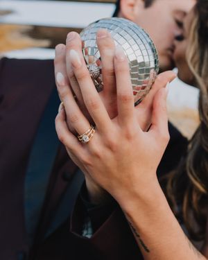 Couple holds a disco ball while kissing, showcasing a gold diamond engagement ring and tattoo on her hand.