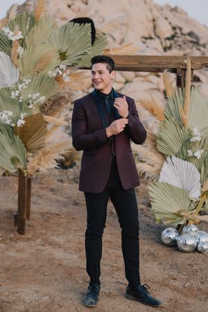 Handsome groom in burgundy velvet blazer adjusts cufflinks before boho desert wedding ceremony backdrop.