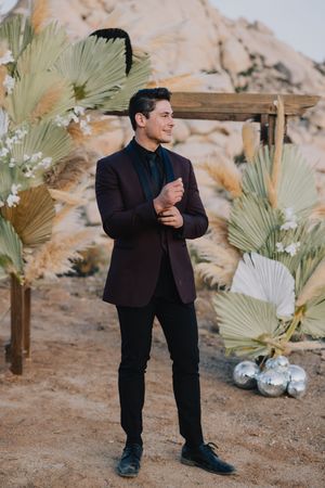 Stylish groom in burgundy blazer standing near desert floral arch with palm leaves.
