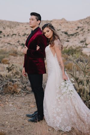 Bride in lace wedding gown and groom in burgundy velvet tuxedo pose back-to-back among desert cacti.