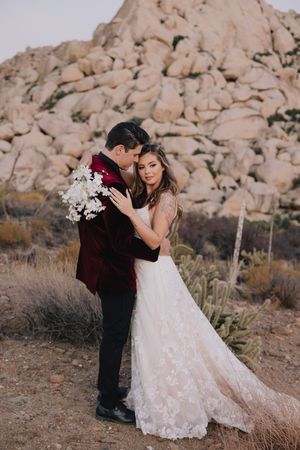 Bride and groom embrace in desert landscape, she in lace gown, he in burgundy velvet suit with floral bouquet.