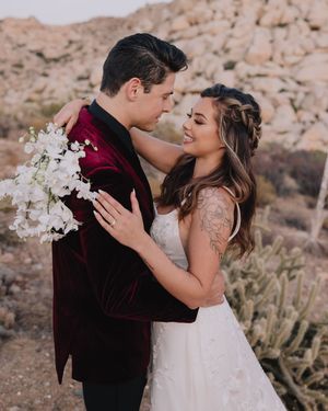 Couple embracing tenderly, groom in red velvet blazer holding white bouquet with rocky backdrop.