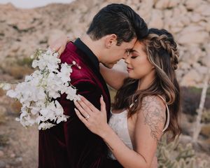 A couple shares an intimate moment in a desert setting, the groom in burgundy velvet holding white orchids.