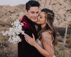 Couple embracing in desert landscape, groom in burgundy velvet suit, bride with tattoo holding white flowers.