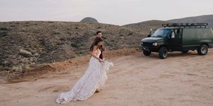 Bride and groom walk hand in hand through sandy desert landscape with vintage green van in background.