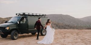 Bride in white gown and groom hold hands beside a green off-road van in a desert landscape.