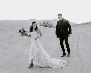 Black and white photo of bride in lace gown with train and groom walking on sandy desert path.
