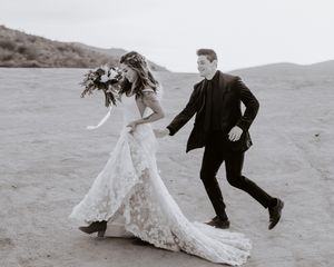 A bride and groom hold hands running through sandy desert landscape, bride holding bouquet, black and white wedding photo.