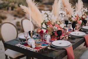Boho outdoor tablescape with pampas grass, pink florals, dark wood table, and colorful runner in a desert setting.