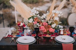 Elegant wedding table setting with pink roses, pampas grass, dark glassware, and colorful floral runner on a rustic wood table.