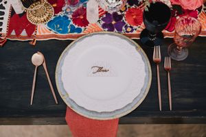 Elegant wedding place setting with white plate, rose gold cutlery, black glassware, and colorful floral table runner.